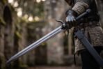 Steel skull sword with blackened horned guard held in a gauntleted hand, scabbard at the hip, ruined stone walls behind