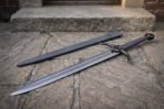 Steel skull sword with chevron etched blade laid on flagstones below stone steps, black leather scabbard parallel to it
