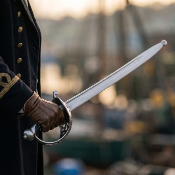 Steel spadroon with a pierced openwork shell guard and curved blade ending in a blunting ball, held by a gloved hand in a dark coat, harbour behind