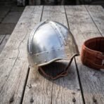 Steel spangenhelm with engraved knotwork browband, brass rivets and leather chin strap on a weathered wooden table