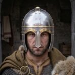 Steel spectacle helmet with a brass brow band, riveted eye guards and domed skull worn by a stern bearded man in fur and chainmail
