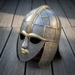 Steel Sutton Hoo helmet with full face mask, brass eyebrows, brass crest band and embossed figure panels lying on grey timber decking