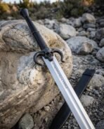 Close up of a steel sword with blackened ram horn skull guard and rune etched blade resting on grey boulders, scabbard tip below