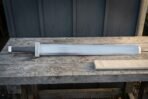 Steel sword with broad fullered blade and knotwork guard laid full length on a weathered bench beside its folded linen scabbard