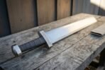 Close up of a steel sword with Celtic knotwork guard, block pommel and dark leather wrapped grip on a weathered wooden bench
