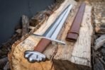 Steel sword with five lobed pommel and etched knotwork fuller resting on a split log, brown leather scabbard alongside