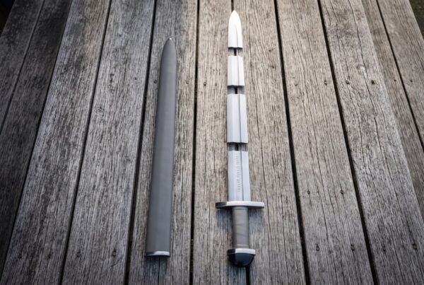 Top down view of a steel sword with grey leather grip laid on decking beside its scabbard, blade split into three pieces