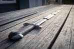 Steel sword with grey leather grip and tea cosy pommel on a weathered wooden deck, blade lying in three separate sections