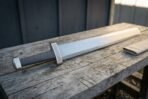 Steel sword with knotwork engraved guard and block pommel laid on a grey wooden bench, folded linen scabbard beside the blade