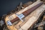 Steel sword with lobed pommel, straight crossguard and brown leather grip laid on a split log beside its brown leather scabbard