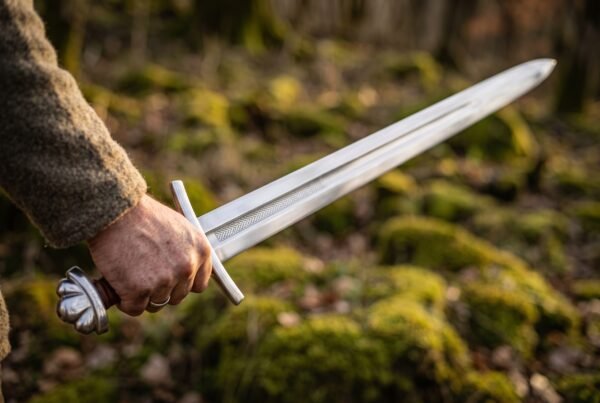 Steel sword with lobed pommel held in hand, etched knotwork running along the fuller, mossy woodland floor behind