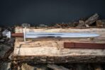 Steel sword with lobed pommel and brown leather grip laid across a split log with its brown leather scabbard in front