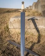 Steel sword with polished brass downswept guard and ribbed pale grip standing upright against a stone block, scabbard at its foot