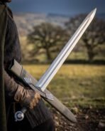 Steel sword with ring pommel drawn from its black scabbard by a gloved hand in a brown cloak, open fields behind