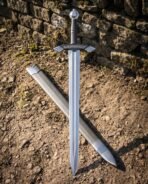 Steel sword with ring pommel and rune cut guard laid on bare earth crossed over its black scabbard, top down view