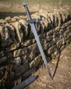 Steel sword with ring pommel and rune cut winged guard leaning against a dry stone wall, black scabbard on the ground