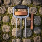 Top down view of a steel three blade knuckle claw on moss covered cobblestones with the tan leather strap laid flat alongside