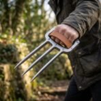 Steel three blade knuckle claw worn on a hand with the tan leather strap over the knuckles, hedgerow out of focus behind