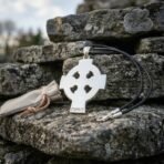 Steel tone Celtic cross pendant with a pierced ring, rune stamped lower arm and black leather cord propped on lichen covered stone
