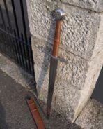 Steel triskele sword with brown leather grip and wheel pommel leaning on a stone gatepost above its tan scabbard