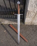 Steel triskele sword with wheel pommel standing point down against iron railings beside its tan leather scabbard