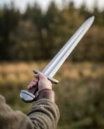 Steel Viking style sword with round pommel and leather grip raised in a hand at arm's length against blurred trees