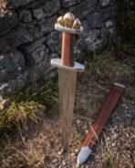 Steel Viking sword with etched valknut ricasso standing point down on gravel by a stone wall with the brown leather scabbard alongside
