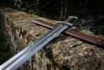 Steel Viking sword with fullered blade and brown leather scabbard resting along the top of a lichen covered stone wall