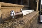 Steel Viking sword with a lobed pommel, brown leather grip and rune row along the blade, resting on sandstone steps close up at the hilt