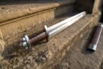 Steel Viking sword with rune engraved blade and lobed pommel laid on sandstone steps with its brown leather scabbard alongside