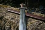 Steel Viking sword with wheel pommel and brown leather grip laid on a mossy stone wall beside its brown leather scabbard