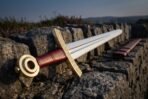 Steel Viking sword with knotwork brass guard, red leather grip and ring pommel leaning on a stone wall with a red scabbard