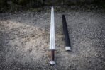 Steel Viking sword with lobed pommel and brown leather grip laid on gravel beside its black leather scabbard, top view
