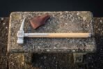 Steel war hammer with Mjolnir engraved head and langets on an ash haft lying on a stone bench beside a leather head cover