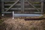 Steel war sword shown full length on bare earth by a wooden gate with its brown leather scabbard alongside