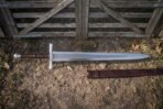 Steel war sword with lattice etched fuller and knotwork guard laid on dry ground in front of a wooden gate