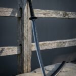 Straight bladed blackened ninjato with a square guard and black cord wrapped handle standing against a weathered timber fence post beside its black lacquered saya