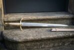 Straight bladed hand forged fantasy arming sword with a brass cross hilt and pale leather grip lying on stone steps beside a matching cream leather scabbard