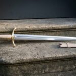 Straight bladed hand forged fantasy arming sword with a brass cross hilt and pale leather grip lying on stone steps beside a matching cream leather scabbard