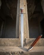 Straight steel Norse sword with disc pommel and squared crossguard standing against a stone pillar beside its russet scabbard