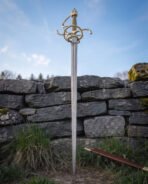 Swept hilt sword planted point down in spring grass below a mossy dry stone wall, brass toned knuckle bow and side rings crowning the slim Damascus blade