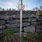 Swept hilt sword standing point down in grass before a dry stone wall and open sky, brass toned knuckle bow and side rings above a wild pattern Damascus blade