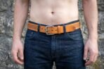 Tan leather belt with Celtic knotwork stamping and brass rivet worn on dark jeans against a stone wall