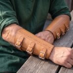 Tan leather rune bracers buckled on a man's forearms as he rests his arms on a weathered wooden table
