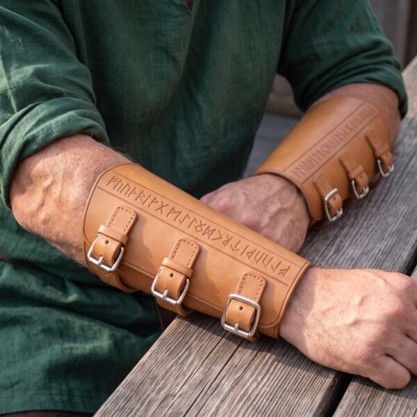 Tan leather rune bracers buckled on a man's forearms as he rests his arms on a weathered wooden table