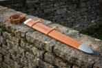 Tan leather rune tooled scabbard with brass throat and steel chape seen at an angle on a stone wall with a coiled belt