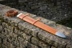 Tan leather sword scabbard tooled with Elder Futhark runes and fitted with a steel chape, laid on a mossy dry stone wall beside its matching buckled belt