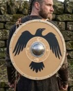 Tan planked round shield with black raven and steel boss slung on the back of a bearded man with braided hair in a grey wool tunic