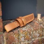 Tan tooled leather knife sheath with Celtic knotwork panel and brass snap strap lying on a brick ledge beside brass rivets