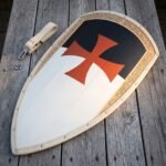 Templar kite shield of planked wood with a black and white field, a red cross pattee and an engraved gilt border strip, on grey decking beside a leather guige.